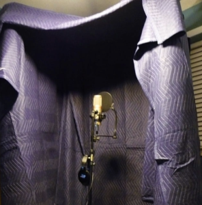 Photograph of a microphone with a blanket draped in a half box shape surrounding it. Original photo by California Dingo: californiadingo.com/blog/tag/diy-vocal-booth/